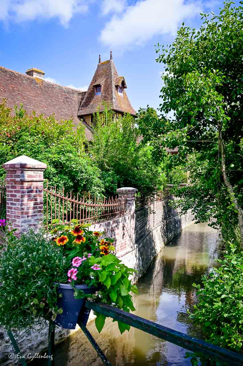 Liten å i Beuvron-En-Auge  med en blomstrande bro och ett stort korsviekshus i bakgrunden